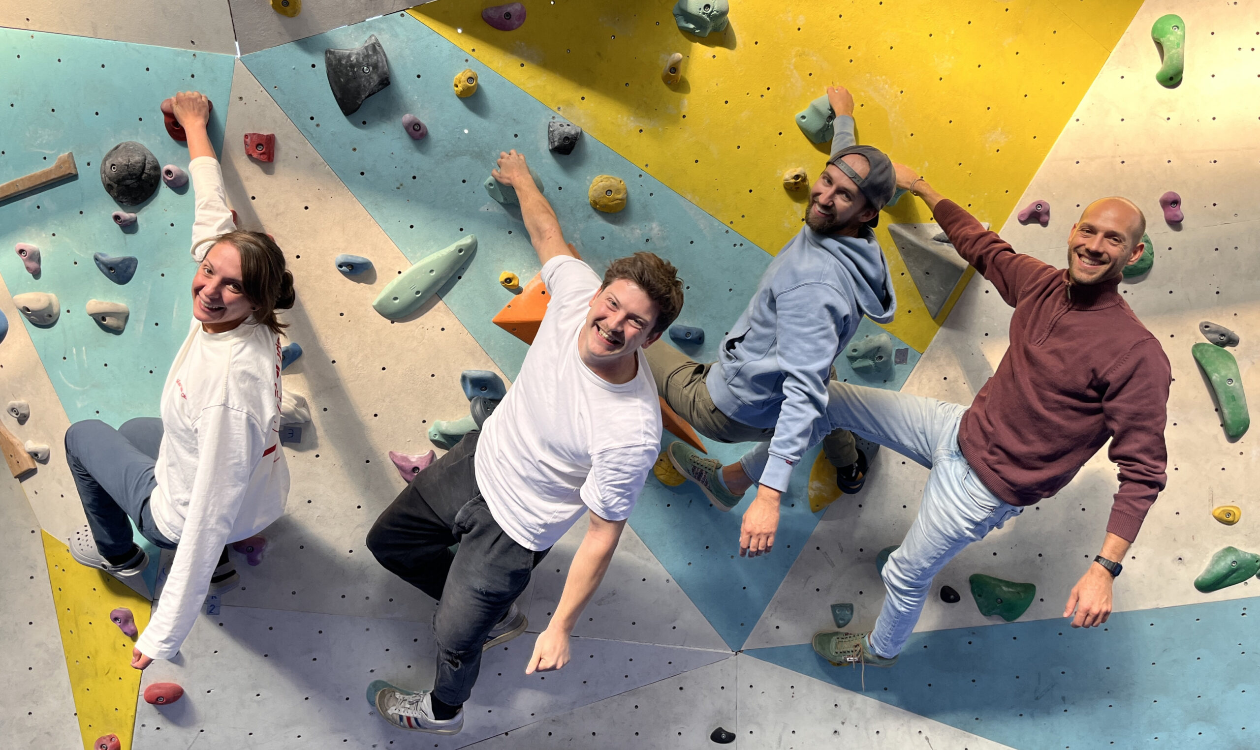 Vier Personen hängen mit einer Hand an einer Boulderwand und lachen in die Kamera